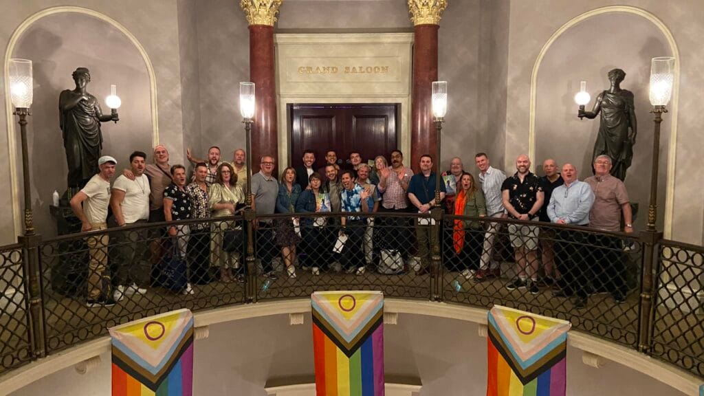 A large group at a queer theatre event on the staircase of a grand London theatre, with Pride flags